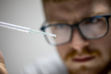 Gözlüklü genç sakallı biyolog yeşil kimyasal reaktörün damlasına bakıyor.