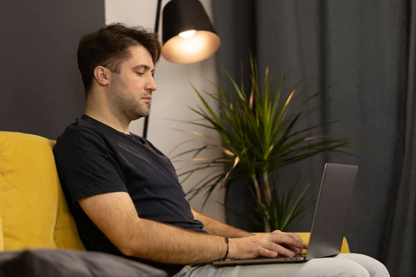 Joven hombre guapo sin afeitar sentado en el sofá y escribiendo en el teclado del ordenador portátil — Foto de Stock