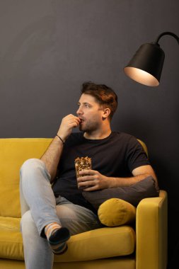 Tıraşsız, sıkılmış bir adam patlamış mısır yer ve rahat sarı kanepeye oturur.