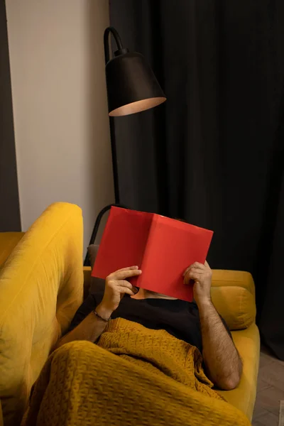 毛むくじゃらの腕男は毛布で覆われた快適な黄色のソファに横たわって本を読む — ストック写真