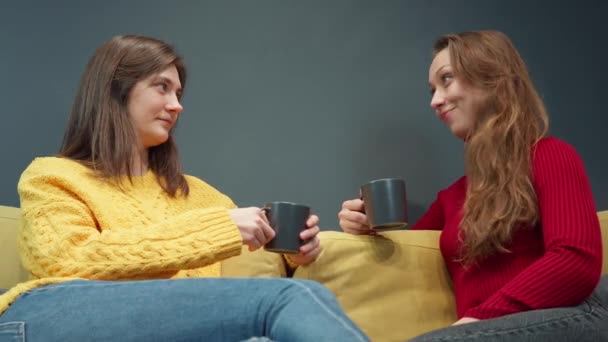 Dos chicas sonrientes discuten chismes sentadas en un acogedor sofá amarillo bebiendo café — Vídeo de stock