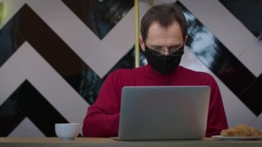 Siyah koruyucu maskeli yakışıklı orta yaşlı adam kafede dizüstü bilgisayarla çalışıyor.