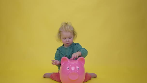 Милая улыбающаяся малышка весело кладет монеты в огромную копилку — стоковое видео