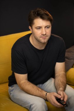 Traş edilmemiş sıkıcı adam TV izlerken uzaktan kumandaya tıklıyor.