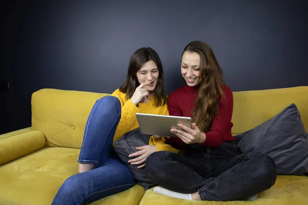 Dos chicas morenas de pelo bastante largo sostienen tableta de chat y mensajería en el sofá — Foto de Stock
