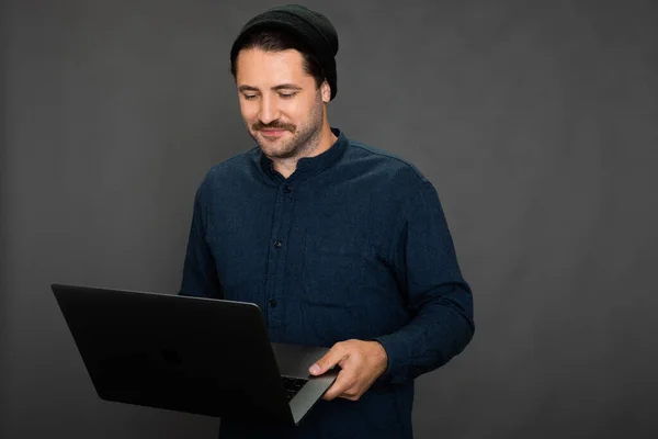 Elegante freelancer masculino mira a la computadora portátil con sonrisa sobre fondo gris, en línea —  Fotos de Stock