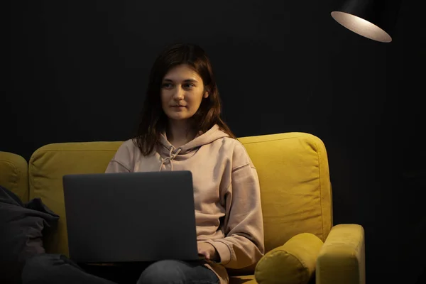 Chica de pelo largo sentado pensativamente en el sofá con el ordenador portátil bajo la lámpara de punto — Foto de Stock