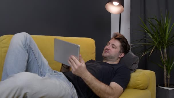 Young unshaven handsome smiling guy messaging online lying on sofa with tablet — Stock Video