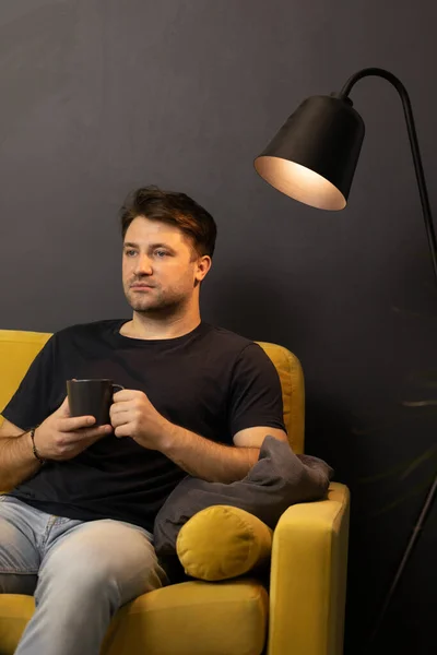 Hombre aburrido sin afeitar con taza de café mirando hacia otro lado sentado en cómodo sofá amarillo — Foto de Stock