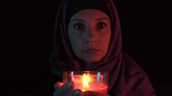 A night portrait of a witch casting spell over a red candle and making witchcraft, she moves a candle close up to the camera and blow out a flame.