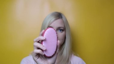 Kırmızı ve pembe saç fırçasını gösteren bir kadın sarı arkaplanda bir kameraya yakın duruyor.