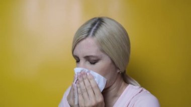 Sarı arka plan, grip ve hastalık konseptine karşı beyaz kağıt mendil kullanan sarışın bir kadın.