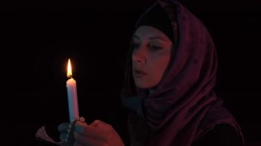 Elinde yanan bir mumla karanlıkta tanrıya dua eden başörtüsü takmış bir kadın.
