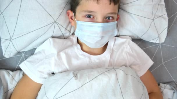 A recovering boy medical protective mask lying in bed and his mother measuring temperature with infrared contactless thermometer, concept of healthy child — Stock Video