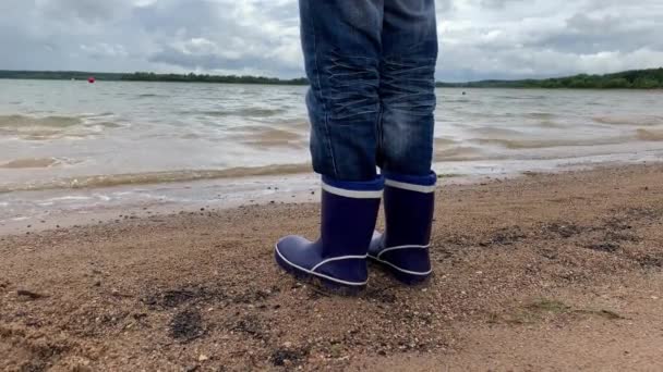 Un niño con botas de goma azul de pie en la arena de la orilla de un lago, se ve en el hermoso paisaje acuático con olas — Vídeo de stock