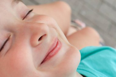 Yazın dışarıda dinlenen bir çocuğun yakın plan yüzü, gözleri kapalı ve gülümsüyor, tatil kavramının tadını çıkarıyor.