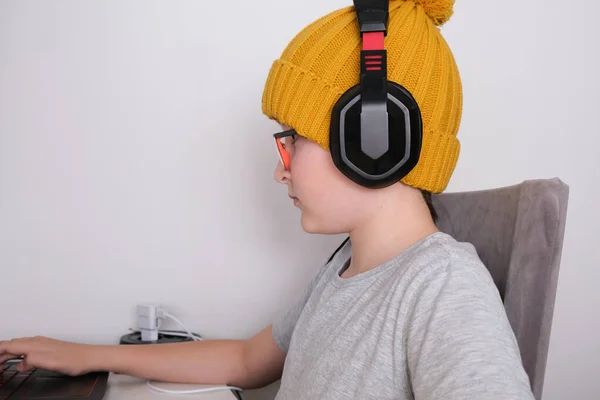 Ένα αγόρι gamer σε ένα μοντέρνο πλεκτό καπέλο και heaphones κάθεται δίπλα στο τραπέζι και παίζει multiplayer παιχνίδι του υπολογιστή στο laptop — Φωτογραφία Αρχείου