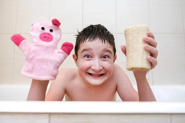 Egy vicces arcú fiú portréja, aki a fürdőkádban ül, és szivacsot és kézimosó ruhát mutat. — Stock Fotó