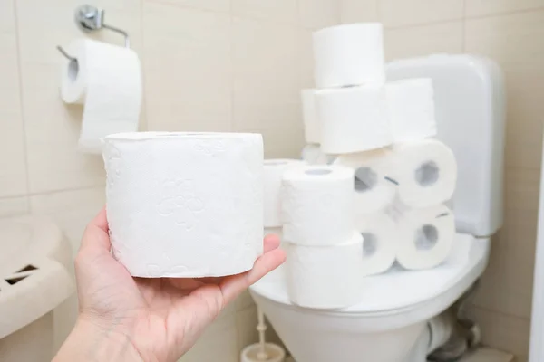 Molti rotoli di carta igienica a casa di accumulatore, l'acquisto di troppi mezzi igienici durante la pandemia, una mano che tiene un rotolo di carta igienica in un bagno — Foto Stock