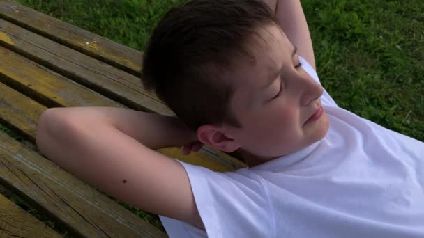 A boy lying on a bench with closed eyes, resting and dreaming outdoors on summer evening — Stock Video