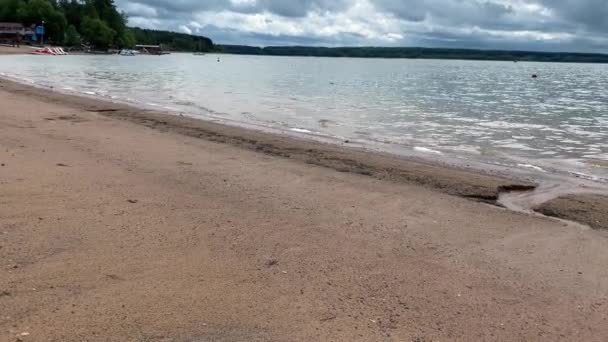 Una riva sabbiosa di un lago con acqua limpida e piccole onde in una giornata nuvolosa in estate — Video Stock