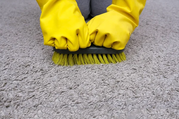 Людина в жовтих гумових світильниках з пензлем в руках прибирає килим вдома, прибирає будинок від пилу — стокове фото