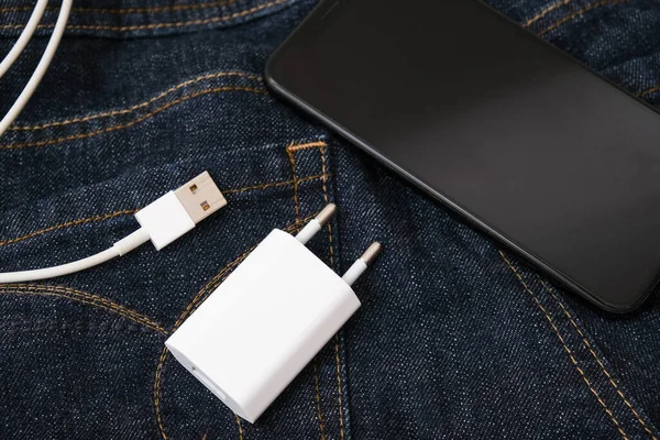 Cargador USB, enchufe de cable y adaptador y un teléfono inteligente en bolsillo azul oscuro jeans, no se olvide del cargador en casa — Foto de Stock