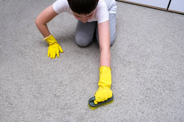 Ein Junge in gelben Gummihandschuhen hilft seiner Mutter beim Teppichputzen mit Pinsel, Hauswirtschaft und Erziehungskonzept — Stockfoto