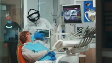 Koruyucu kıyafetli stomatolog, tablet kullanarak diş röntgenini inceliyor.