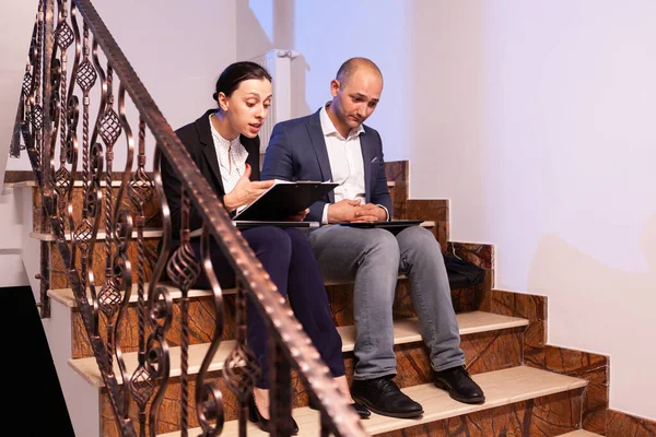 Worried tired businessman doing overtime at night on job diffuclt deadline with coworker sitting on stairs. — Stock Photo, Image