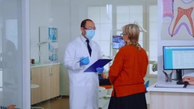 Maskeli ortodontist bekleme salonunda yaşlı bir kadınla konuşuyor.