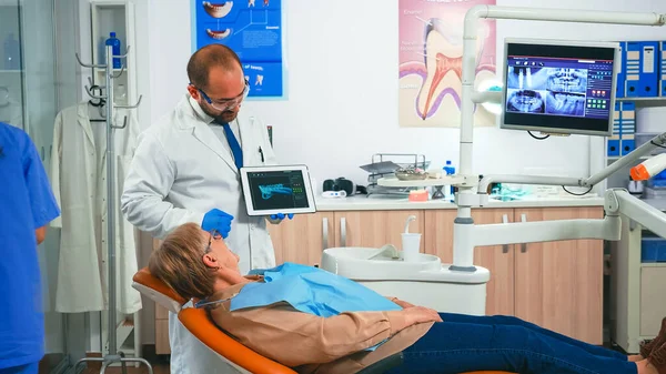 Vrouw zittend op tandartsstoel luisterend arts kijkend op tablet — Stockfoto