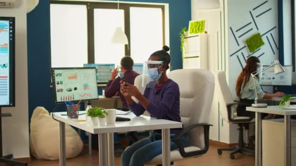 Mujer negra navegando en el teléfono inteligente sentado en una nueva oficina normal — Vídeo de stock