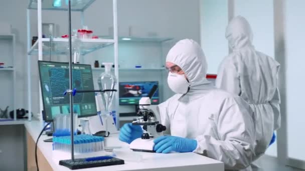 Científico en traje ppe trabajando en laboratorio usando microscopio moderno — Vídeos de Stock