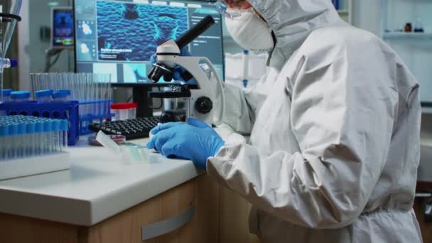 Científico en traje de protección mirando a través de un microscopio — Vídeos de Stock