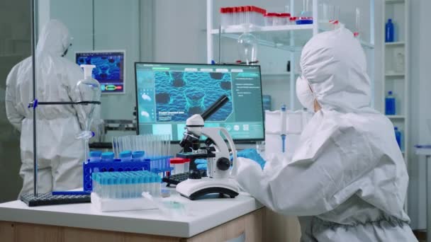 Chemik w kombinezonie ochronnym wpisujący na komputerze i analizujący próbkę wirusa — Wideo stockowe