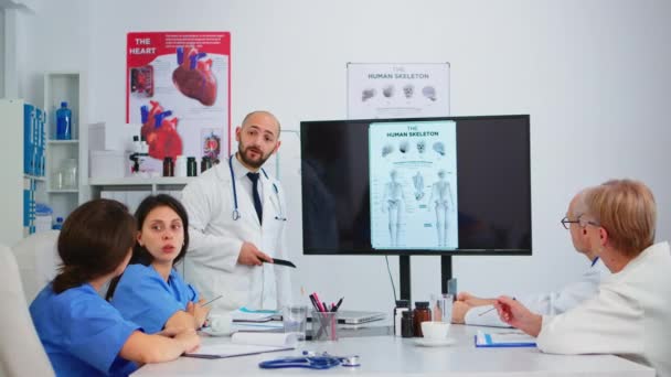 Опытный человек врач анализирует изображение скелета человека с коллегами — стоковое видео