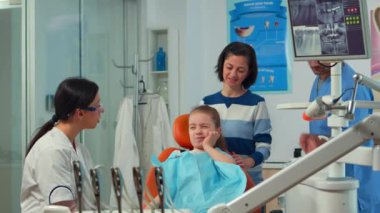 Çocuk, dişçi annesiyle konuşurken dişini işaret etmek için parmağını kullanıyor.