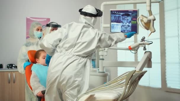 Dokter gigi dalam setelan ppe menunjuk pada monitor gigi digital menjelaskan x-ray kepada ibu — Stok Video