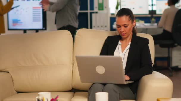 Mujer de negocios hispana sonriendo a la cámara sentada en el sofá escribiendo en la computadora — Vídeos de Stock