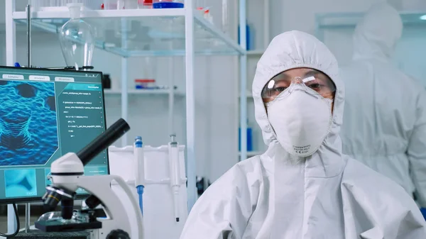 Микробиолог, сидящий в лаборатории в костюме ppe, смотрит в камеру — стоковое фото