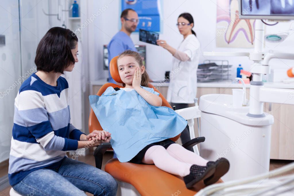 Little girl in dentist office showing parent where tooth hurts touching face