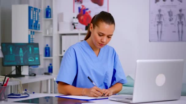 Arzthelferin tippt auf Laptop und macht sich Notizen auf Klemmbrett — Stockvideo