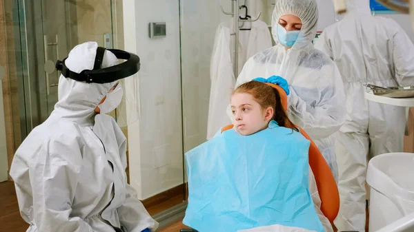 Little girl with dental bib and ppe suit listening pediatric dentist in coverall