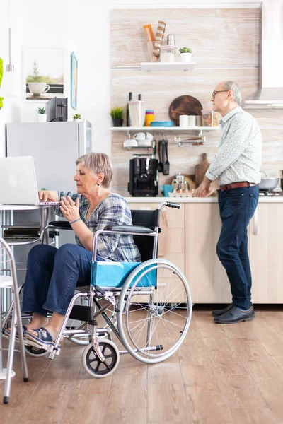 坐在轮椅上的残疾老年妇女在厨房的笔记本电脑旁工作 — 图库照片