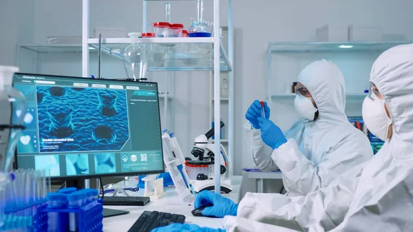 Доктор в костюме ppe работает за компьютером, в то время как химик использует микроскоп — стоковое фото