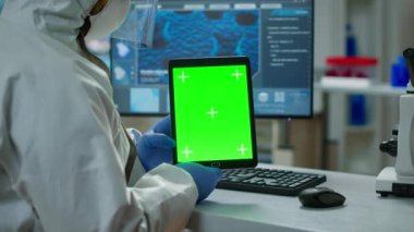 Koruyucu giysili kimyager elinde yeşil bir tablet tutuyor.