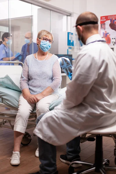 Старша жінка в масці для обличчя в лікарні, слухаючи діагноз лікаря — стокове фото