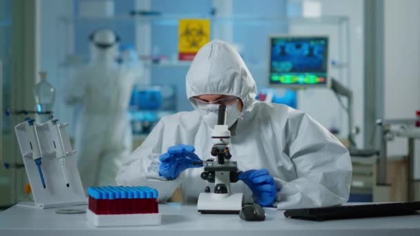 Naukowiec w garniturze ppe korekty i patrząc przez mikroskop laboratoryjny — Wideo stockowe