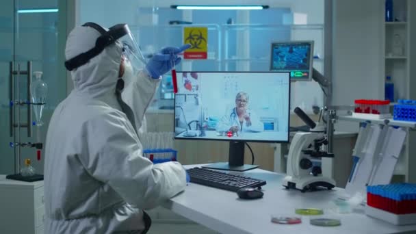 Tecnico di laboratorio in tuta protettiva che tiene la provetta e parla in videochiamata con il medico — Video Stock